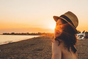 Das Bild zeigt eine junge Frau mit Sonnenhut am Strand in der Abendsonne stehen.