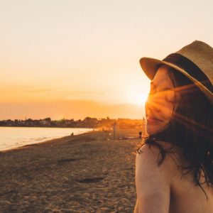 Das Bild zeigt eine junge Frau mit Sonnenhut am Strand in der Abendsonne stehen.